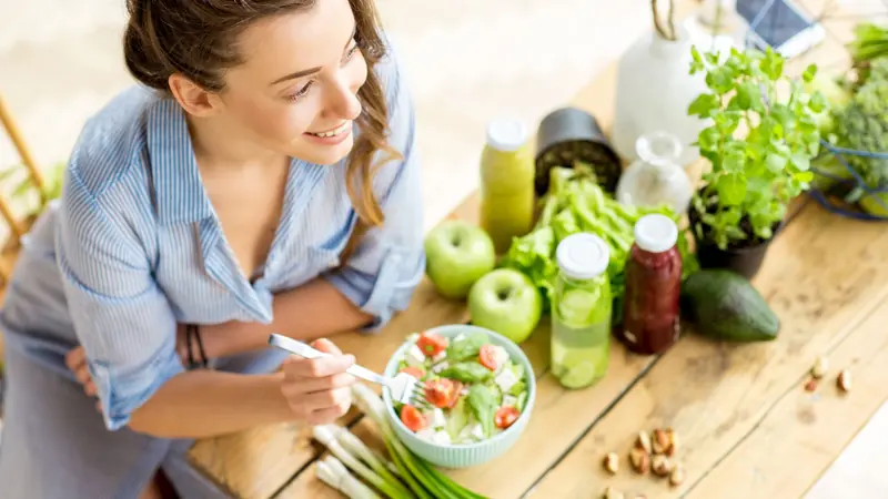 A woman smiling while eating a fresh salad at a kitchen table with vegetables and herbs